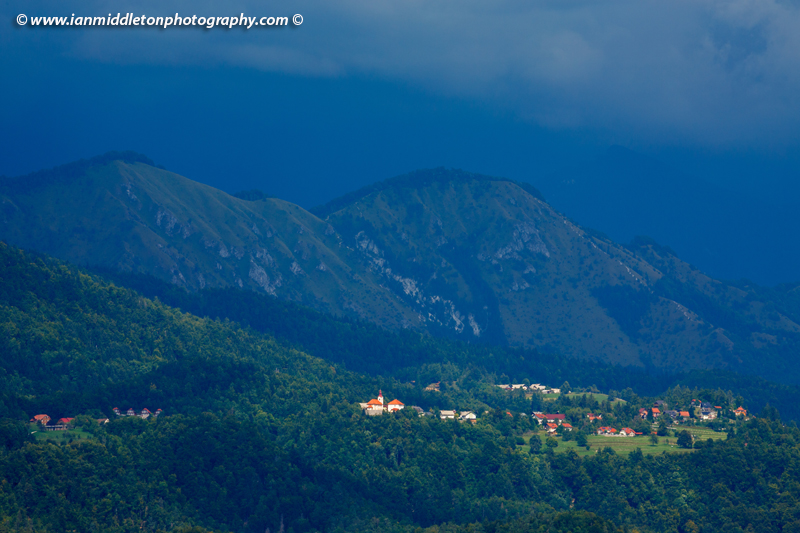 The small village of Šenturška Gora spot lit as a summer storm passes over. The village sits in the footihills of the Kamnik Alps, Gorenjska Slovenia. The photo was taken from Brnik, near the airport.
