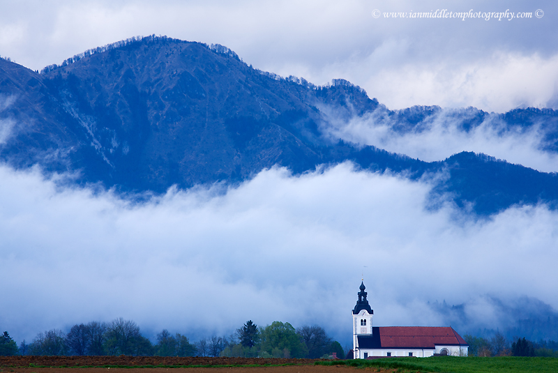 mountaintop of the Kamnik Alps appearing through the rain clouds after an evening storm with the church of Saint Peter sitting in its shadow. This complex also contains a castle, a baroque church, a parsonage, a cemetery and a fairly large stable.