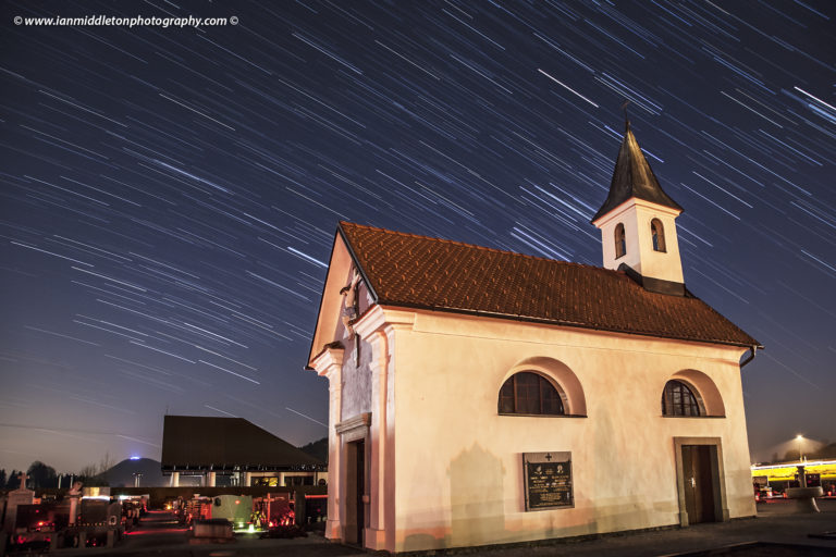 Star trails behind a small chapel in Vodice, Gorenjska, Slovenia. The brightest stars around the church spire are from the Orion constellation. This small graveyard is also lit by candles on the graves, which is a common practice here in Slovenia. This image was captured just as the moon was waning at 96.4% and just rising to the east, which, along with cars and streetlights, helped to nicely illuminate the chapel and graveyard.