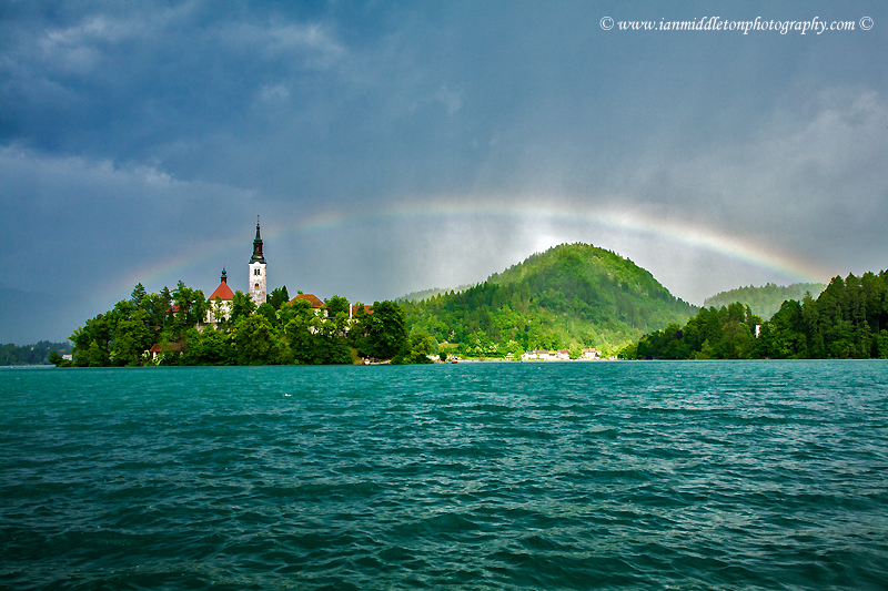 Beautiful light across to the beautiful Lake Bleds island church as a storm blows in and produces a rainbow right over the church and surrounding hills, Slovenia.