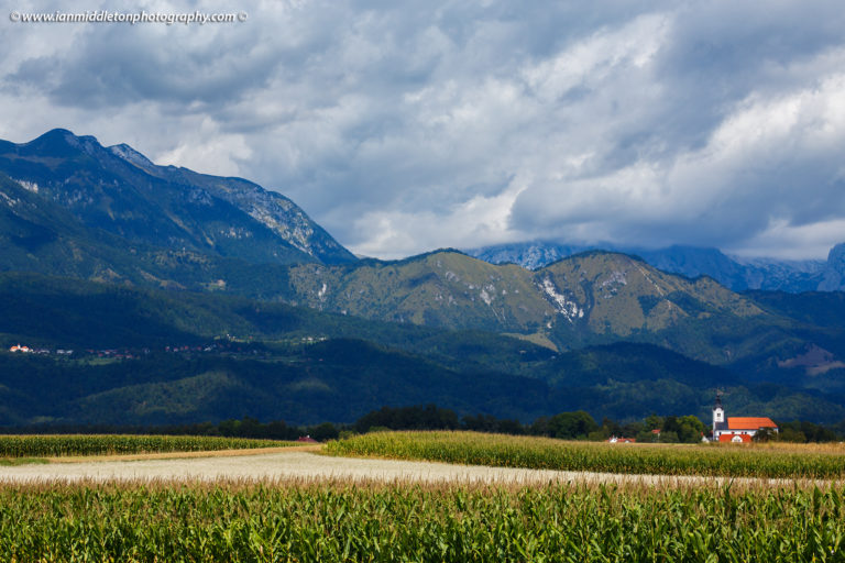 Mountaintop of the Kamnik Alps appearing through the clouds with the church of Saint Peter sitting in its shadow. This complex also contains a castle, a baroque church, a parsonage, a cemetery and a fairly large stable.