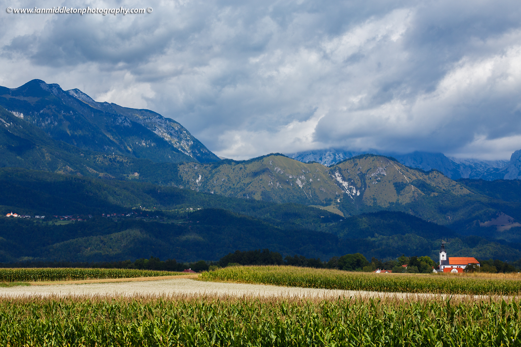 Mountaintop of the Kamnik Alps appearing through the clouds with the church of Saint Peter sitting in its shadow. This complex also contains a castle, a baroque church, a parsonage, a cemetery and a fairly large stable.