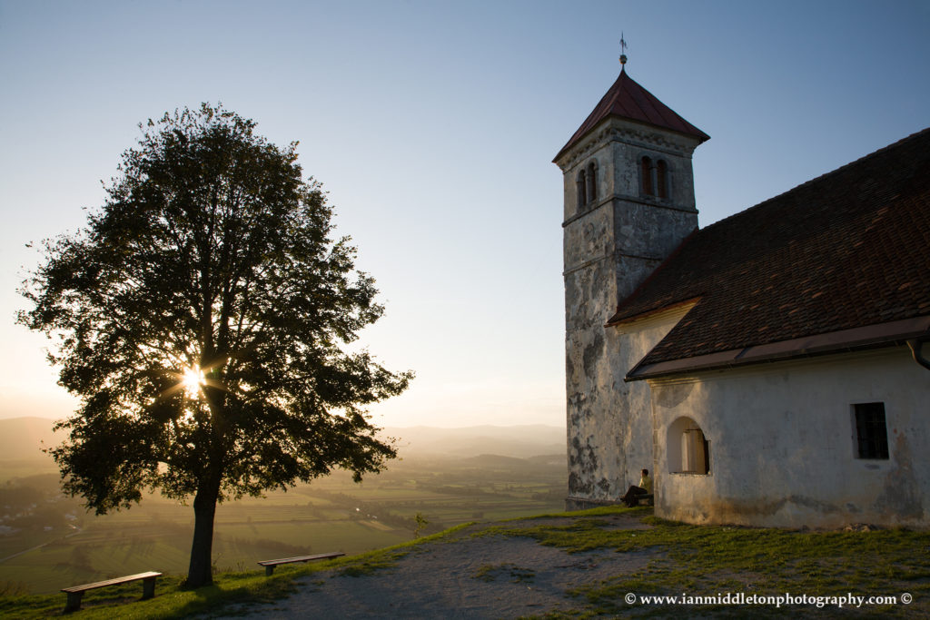 Soft light falls over the church of Saint Anna (Sveta Ana) as the sun falls down behind a nearby tree causing a sunburst through the leaves and branches. Sveta Ana is perched upon an exposed hill overlooking the Ljubljansko Barje (Ljubljana marsh) near the village of Preserje. The views from this hill are spectacular and many locals seem to come here for the evening.