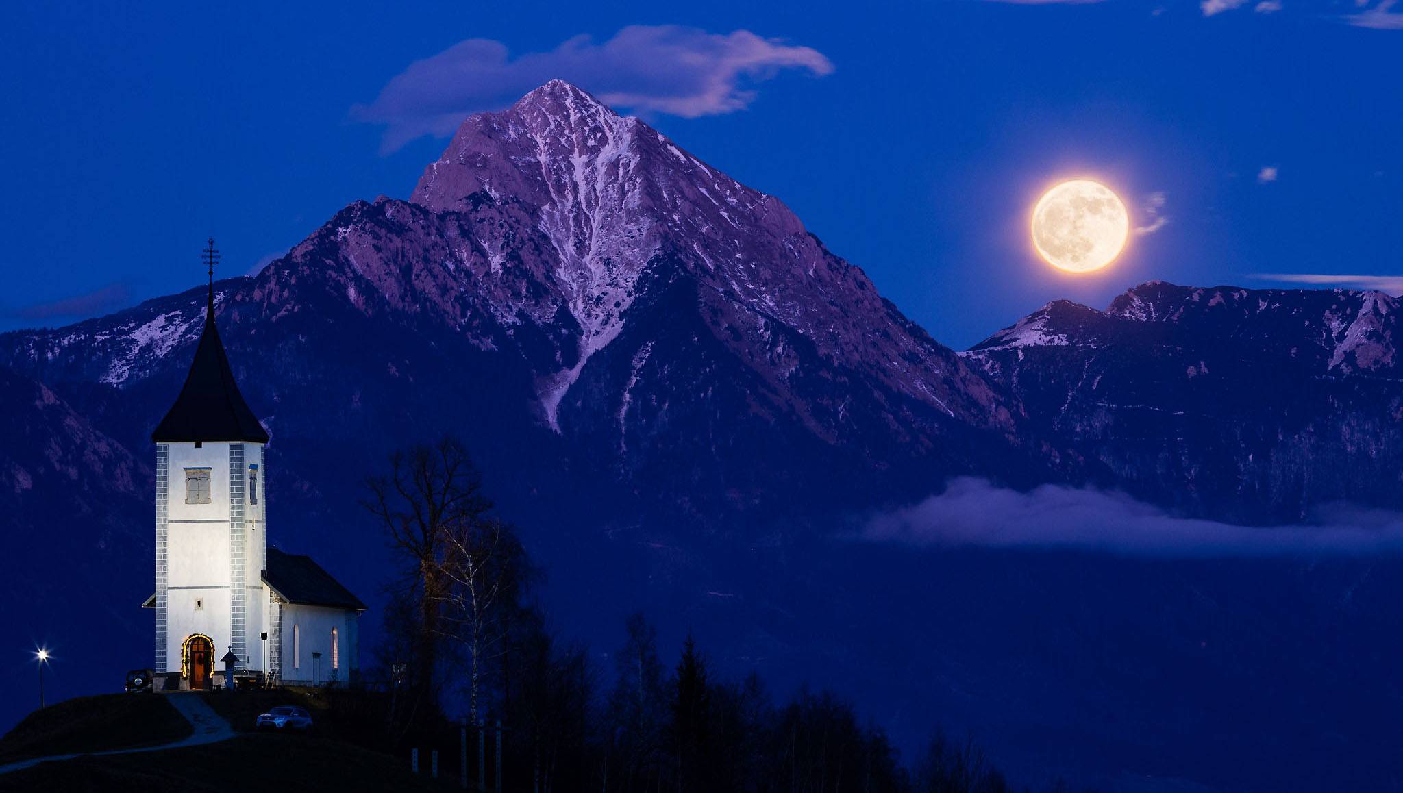 Full moon rising at dusk over Jamnik church of Saints Primus and Felician, perched on a hill on the Jelovica Plateau with the kamnik alps and Storzic mountain in the background, Slovenia.