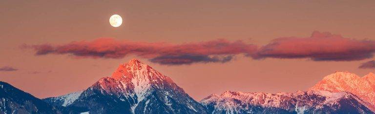 Full moon rising at sunset over Jamnik church of Saints Primus and Felician, perched on a hill on the Jelovica Plateau with the kamnik alps and Storzic mountain in the background, Slovenia.