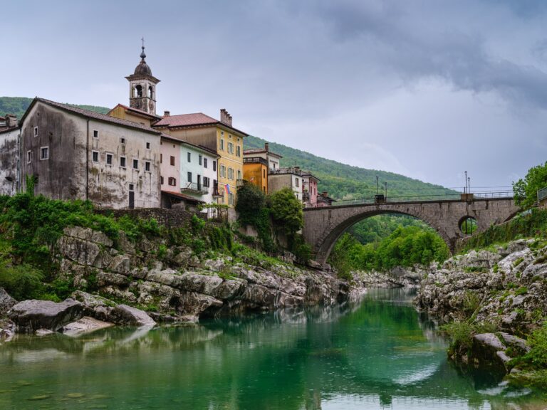 KANAL OB SOČI, SLOVENIA - CIRCA MAY 2024: A picturesque view of the historic village of Kanal ob Soči, with its charming colorful houses perched on the rocky banks of the Soča River. The scene is dominated by the old stone bridge and the church tower, set against a backdrop of lush green hills and a cloudy sky. This image captures the serene beauty and cultural heritage of this Slovenian village.