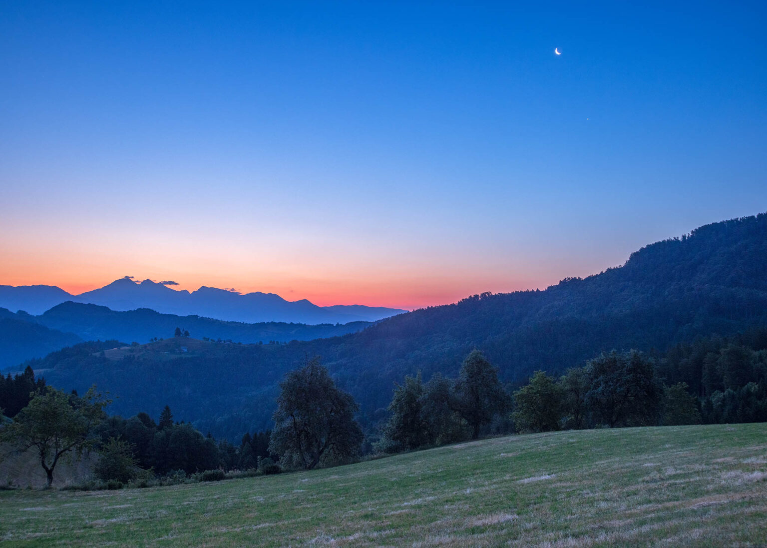 Dawn view on the day after the Summer Solstice from Rantovše hill across to Sveti Tomaz nad Praprotnim (church of Saint Thomas), the Kamnik Alps, with the waning crescent Moon and Venus beside them, Slovenia.