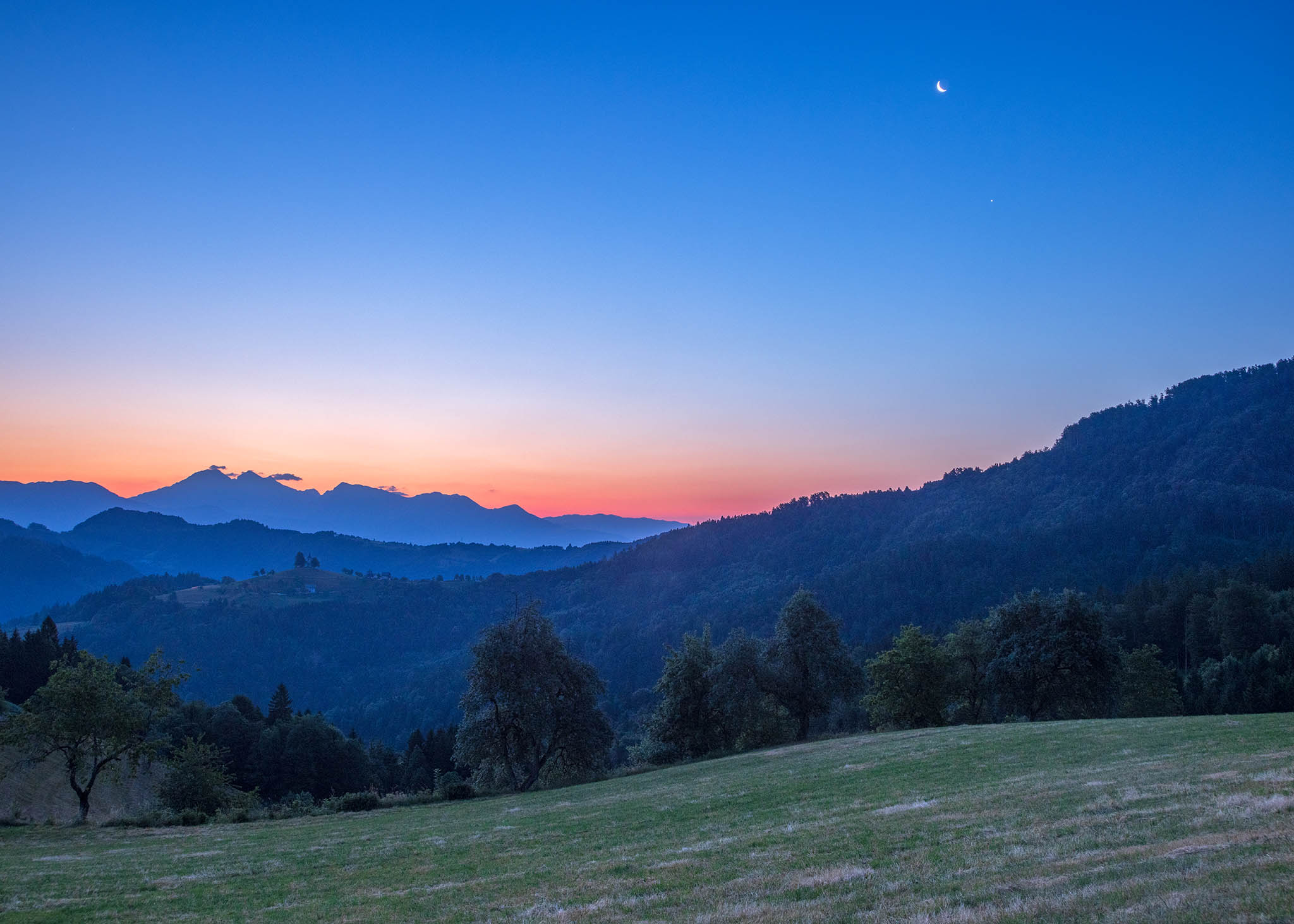 Dawn view on the day after the Summer Solstice from Rantovše hill across to Sveti Tomaz nad Praprotnim (church of Saint Thomas), the Kamnik Alps, with the waning crescent Moon and Venus beside them, Slovenia.