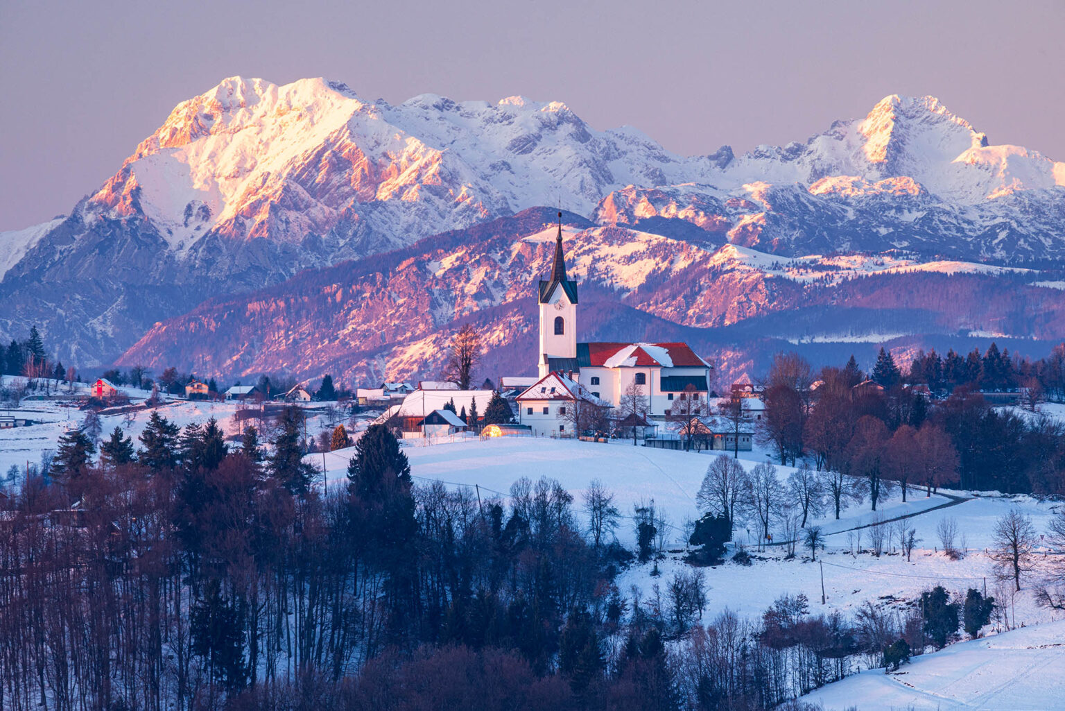 Winter sunset over the church of Saint Marjeta in Prezganje in the Jance hills to the east of Ljubljana, Slovenia. The snow covered Kamnik Alps form a beautiful backdrop.