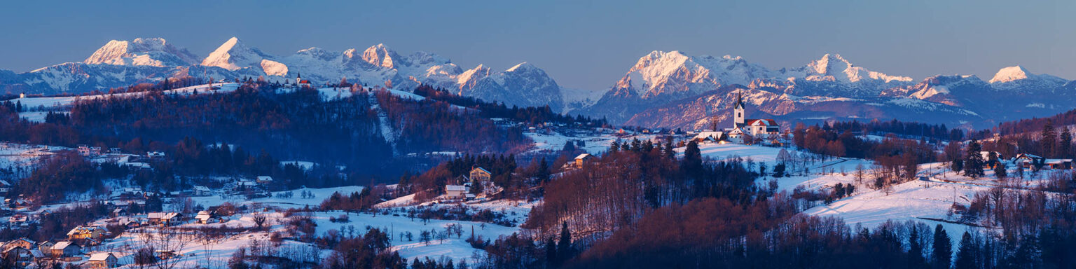 Stitched panorama of winter sunset over the church of Saint Marjeta in Prezganje in the Jance hills to the east of Ljubljana, Slovenia. The snow covered Kamnik Alps form a beautiful backdrop.